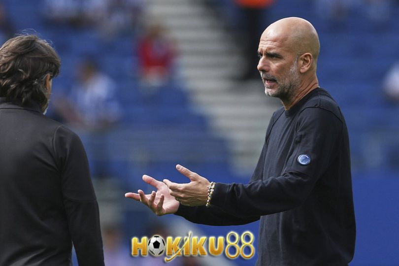 "Pelatih sepak bola memberikan instruksi dan berdiskusi di pinggir lapangan dengan ekspresi serius, mengenakan sweater hitam, latar belakang stadion biru."